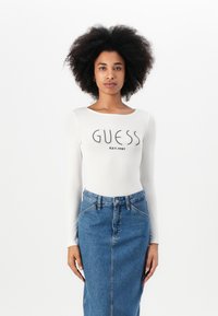 Valkoinen pitkähiha bodysuit, jossa on "GUESS EST.1981" logo, yhdistettynä siniseen farkku-midi-hameeseen, jossa on etunappilista ja taskuja.