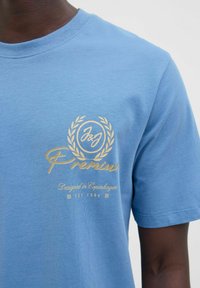 Tricou din bumbac de culoare albastru deschis, cu decolteu rotund, având un logo și un design cu text imprimat în aur pe piept, textură netedă.