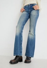 Jeans de campana azules con un ligero desgaste, leves roturas y diseño de cinco bolsillos, combinados con botines negros brillantes.
