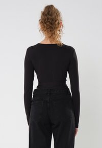 Frau mit lockigem Haar, die ein schwarzes Langarmoberteil und hochtaillierte schwarze Jeans trägt, steht vor einem schlichten hellen Hintergrund.