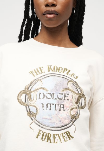 Γυναίκα που φοράει λευκό φούτερ με χρυστό σχέδιο φιδιού και το κείμενο "THE KOOPLES DOLCE UTTA FOREVER" στο μπροστινό μέρος, μπροστά από απλό φόντο.