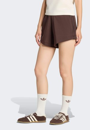Person iført brune shorts, hvide crew-strømper med logo og brune sneakers med hvide striber, står på en lysegrå baggrund.