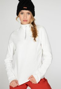 Maglia bianca in pile a maniche lunghe con zip fino a metà, caratterizzata da un logo ricamato e un collo alto. Abbinata a pantaloni rossi vivaci.
