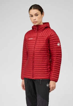 Jeune femme portant une veste matelassée rouge à capuche et un pantalon noir avec des logos de marques outdoor sur un fond gris uni.