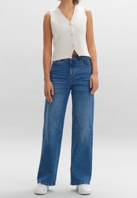 Kvinna som bär ett ärmlöst vitt knäppt väst, högmidjade bredbeniga blå jeans och vita sneakers mot en enkel bakgrund.