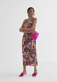 Vestido sin mangas en vibrantes patrones abstractos de púrpura, verde y beige. Combinado con un bolso fucsia y tacones a juego, con volantes.