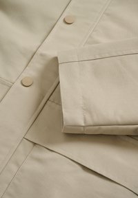 Bež tkanina v bližnjem posnetku z vidnimi šivi, žepkom z zavesico in okroglimi gumbi. Material ima gladko, rahlo teksturirano površino.