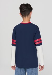 Granatowa koszulka z krótkim rękawem, z czerwonymi paskami na rękawach i czerwonym kołnierzykiem. Nosi się ją na białej koszuli z długim rękawem.