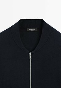 Navy stickad bomberjacka med ribbad krage och en silvrig dragkedja. Den inre etiketten visar "Massimo Dutti." Mjuk textur, avslappnad design.