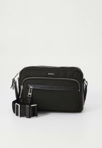 Juoda BOSS rankinė su keliomis užtrauktukais ir reguliuojama dirželiu ant baltos paprastos fono.