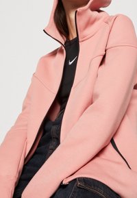 Rosa Kapuzenpullover mit Reißverschluss und hohem Kragen, aus weichem Material gefertigt. Er verfügt über schwarze Reißverschlüsse und ein tailliertes Design mit Nähten an Vorderseite und Ärmeln.