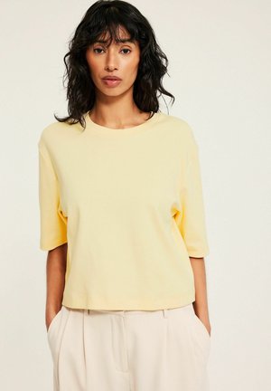 Vrouw met donker golvend haar, gekleed in een lichtgele shirt met korte mouwen en crèmekleurige, hooggetailleerde broek, met handen in de zakken, tegen een neutrale achtergrond.