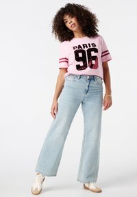 Lichtroze t-shirt met "PARIS 96" in pailletten en korte mouwen, gecombineerd met wijde lichtblauwe jeans en witte sneakers.
