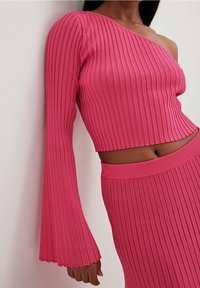 Vaaleanpunainen ribattu neulecrop-toppi, jossa on yksi pitkä hiha ja toinen olka paljaana, yhdistettynä samasta materiaalista valmistettuun ribattuun hameeseen. Kankaan teksturointi näkyy hyvin.