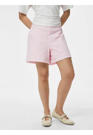 Kvinne som har på seg lyserosa broderte shorts, hvit topp og beige casual sko, står med hendene bak ryggen.