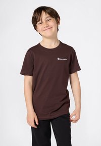 Ragazzo giovane sorridente che indossa una maglietta marrone Champion e pantaloni neri, in piedi con una postura rilassata contro uno sfondo semplice.