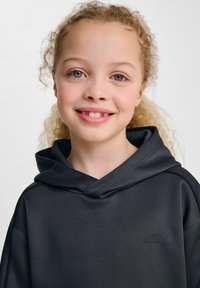 Svart hoodie med hög krage, tillverkad av mjukt tyg. Har en logotyp på framsidan och en avslappnad passform, lämplig för vardagsbruk.