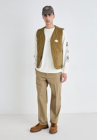 Chaleco acolchado oliva sobre una camisa de manga larga blanca, combinado con pantalones caqui y botas marrones con cordones. Gorra gris con detalle de logo.