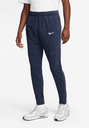 Person wearing navy blue Nike sweatpants with white logo and white sneakers, standing against a plain background.