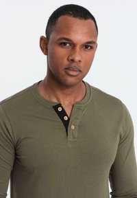 Camicia a maniche lunghe verde oliva con una patta dei bottoni nera, dotata di due bottoni in legno e un tessuto morbido e testurizzato.