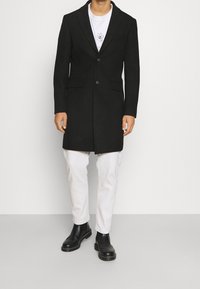 Cappotto di lana nero con spalle strutturate, chiusura a un bottone, due tasche frontali, indossato sopra una maglietta bianca e abbinato a stivali neri.