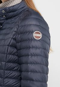 Woman wearing a navy blue quilted jacket with a snap-button collar and a round Colmar logo patch on the upper sleeve.