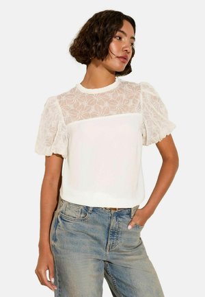Blusa bianca con yoke in pizzo con motivi floreali, maniche corte a sbuffo e corpo uniforme. Abbinata a jeans azzurri chiari.