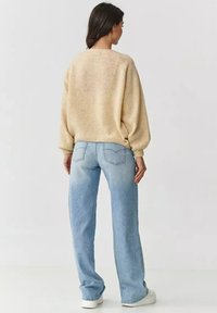 Beżowy sweter dzianinowy o luźnym kroju, zestawiony z jasnoniebieskimi spodniami o prostym kroju. Gładka tekstura i ściągacze na rękawach. Widoczny biały obuwie.