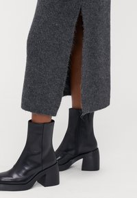 Μαύρες δερμάτινες μπότες έως τον αστράγαλο με χοντρό τετράγωνο τακούνι, συνδυασμένες με γκρι πλεκτό ένδυμα που διαθέτει πλαινές σχισμές και ραφή ριμπ.