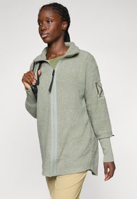 Fleece bunda svetlozelenej farby, s predným zipsom, vysokým golierom a štruktúrovanou tkaninou. Dlhé rukávy s akcentovanými detailmi na manžetách.