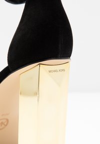 Svart mocka högklackad sandal med en geometrisk guldig klack, prydd med det ingraverade "MICHAEL KORS"-logot. 