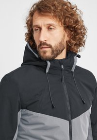 Svart og grå zip-up jakke med hette, med et kontrasterende chevron-mønster, snøring for justering og en jevn tekstur.