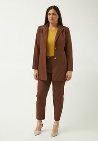 Blazer marrone su misura con rever a punta, abbinato a un top giallo senape e pantaloni coordinati. Le scarpe hanno la punta appuntita e dettagli in metallo sulla caviglia.