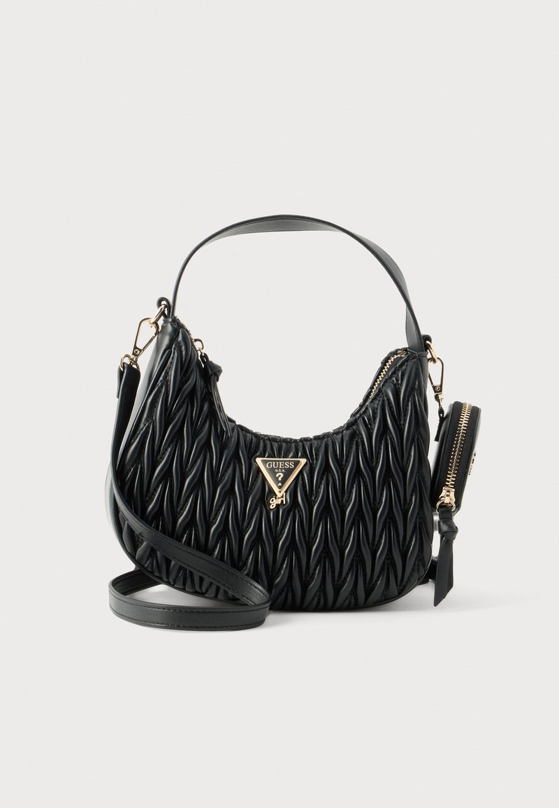 Μαύρη υφασμάτινη τσάντα Guess με χρυσό λογότυπο, κοντή λαβή, μακρύ λουράκι χιαστί και μικρό τσεπάκι με φερμουάρ προσαρτημένο σε απλό φόντο.