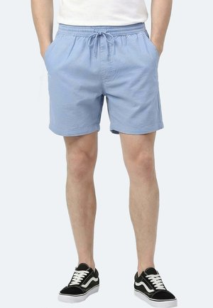 Mann trägt hellblaue Shorts mit Kordelzug, die Hände in den Taschen, und schwarze Sneakers mit weißen Schnürsenkeln und Sohlen, steht vor einfarbigem Hintergrund.