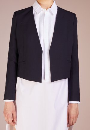 Frau trägt einen eng anliegenden schwarzen kurzen Blazer über einer weißen plissierten Bluse mit Knöpfen und einem weißen Rock vor neutralem Hintergrund.