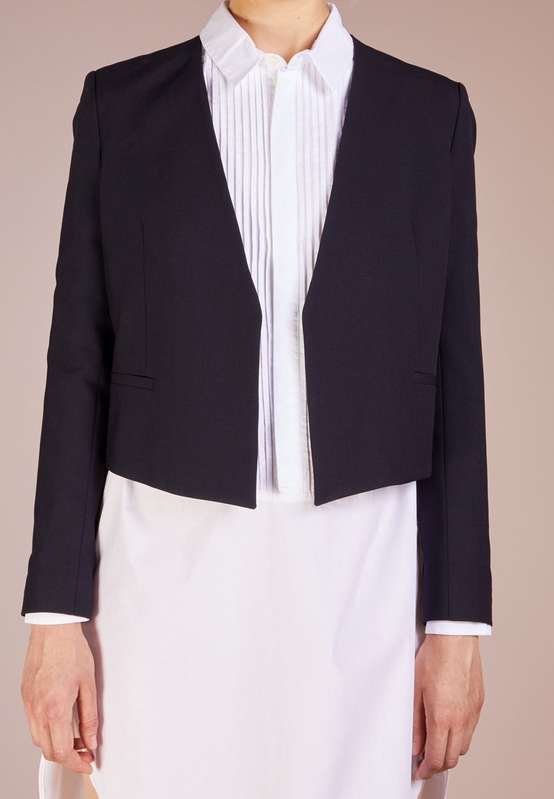 Femme portant un blazer noir ajusté et court sur une chemise blanche plissée boutonnée et une jupe blanche sur un fond uni.
