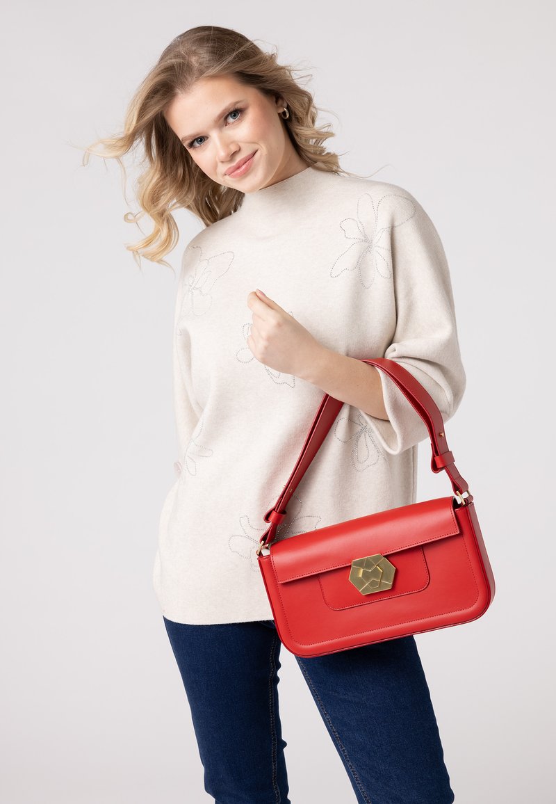 Borsa a tracolla rossa in pelle con una chiusura esagonale dorata, caratterizzata da una forma strutturata e una tracolla removibile. Il modello indossa un top beige e jeans blu.