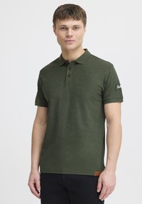 Mørkegrøn polo-shirt med tekstureret stof, tre brune knapper og et logo-detalje på ærmet. Korte ærmer og klassisk krave-design.