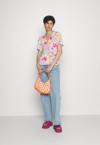 Camisa de flores com corte envolvente em tons pastel, combinada com jeans azul claro, uma vibrante bolsa de rede laranja e sandálias roxas de plataforma.