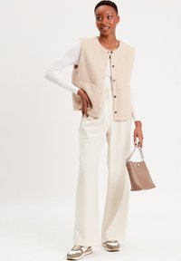 Gilet beige texturé avec poches avant, orné de boutons marron. Associé à un pantalon large crème et à un sac à main marron avec une bandoulière en chaîne.
