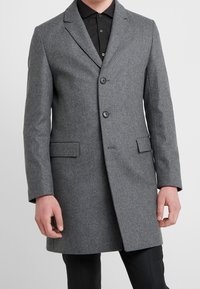 Homme portant un manteau gris en laine, boutonné simple, longueur genou, par-dessus une chemise noire à boutons et un pantalon noir.
