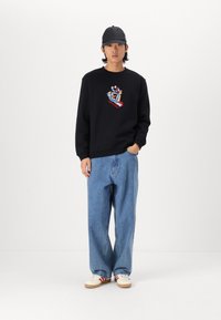 Felpa nera con grafica colorata, jeans blu larghi e sneaker bianche con dettagli rossi, completati da un cappellino nero.