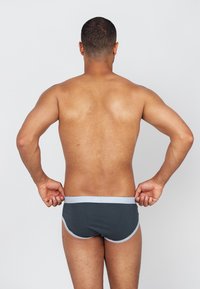 Dunkelgraue Unterhosen aus Baumwolle mit einem hellgrauen elastischen Bund und geschwungenen Seitennähten, gezeigt auf einem nackten Oberkörper von hinten.
