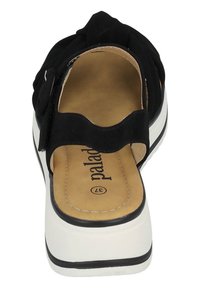 Zapato negro tipo slip-on con parte superior de tela suave, correas anchas y plantilla acolchada. Presenta una suela de plataforma blanca con ribete negro.