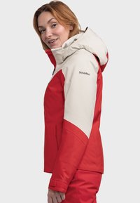 Red and beige waterproof jacket with a hood, side pockets, and an adjustable fit. Features a logo on the sleeve and a smooth texture.
