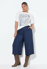 Witte t-shirt met zwart abstract ontwerp, gecombineerd met marineblauwe wijde broek en bruine cowboylaarzen. Model heeft kort haar en staat in een ontspannen houding.