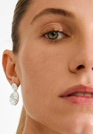 Hõbedast kõrvarõngad spiraaldisainiga, millel on ümmargune ripatsikujuline ja tekstureeritud detail, eksponeeritud lähedalt vaadatava näo kõrval.