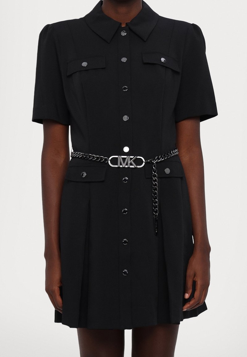 Robe noire à manches courtes avec un col, boutons devant, poches poitrine et une ceinture en chaîne avec un logo. Texture de tissu lisse.