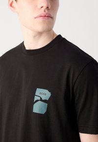 Schwarzes Baumwoll-T-Shirt mit einem blauen grafischen Design auf der linken Seite, in das das Wort "BOSS" integriert ist. Rundhalsausschnitt.